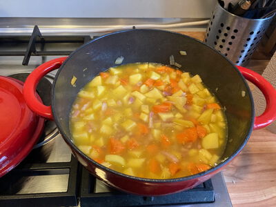 Eine rote Cocotte (gusseiserner Topf) steht auf einem Gasherd. Darin ist ein Gemüseeintopf zu sehen, mit Karotten, Speck, Zwiebeln und Kartoffeln.