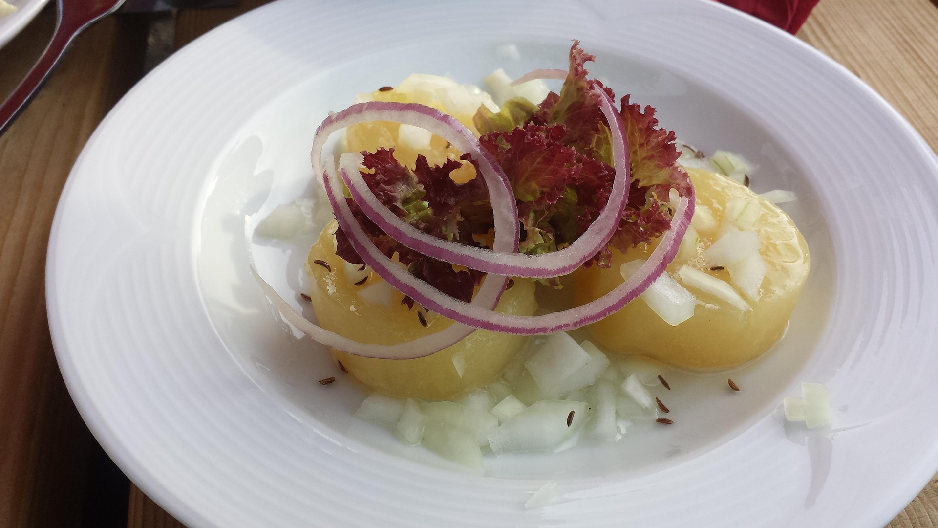 Auf einem Holztisch steht ein weißer Teller mit drei runden Handkäs-Stücken. Darauf liegt ein Salatblatt und rote Zwiebelringe, darum herum die Zwiebelvinaigrette als ""Musik"" und einige Kümmelsamen.  Bild von Jochen Gerlach via Pixabay
